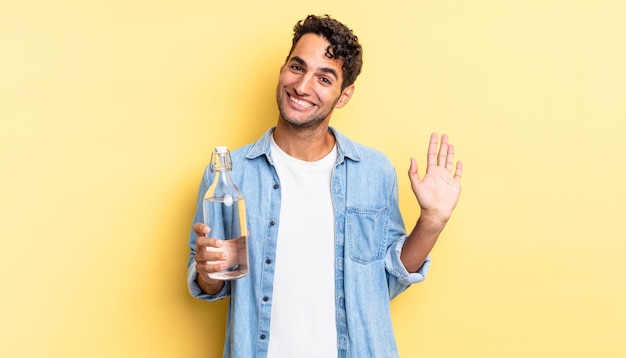 Bel homme hispanique souriant joyeusement, agitant la main, vous accueillant et vous saluant. concept de bouteille d'eau