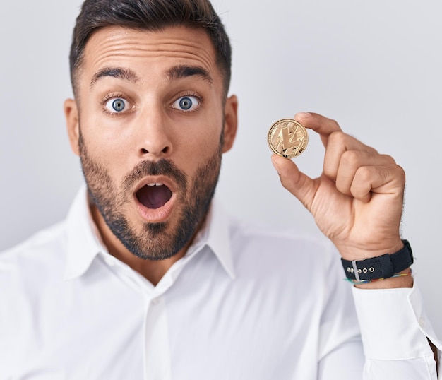Bel homme hispanique tenant une pièce de monnaie crypto-monnaie litecoin effrayé et étonné avec la bouche ouverte pour la surprise, le visage d'incrédulité
