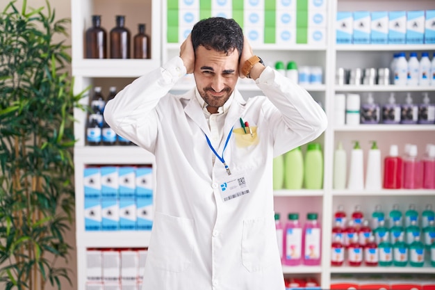 Bel homme hispanique travaillant à la pharmacie souffrant de maux de tête désespérés et stressés à cause de la douleur et de la migraine. mains sur la tête.