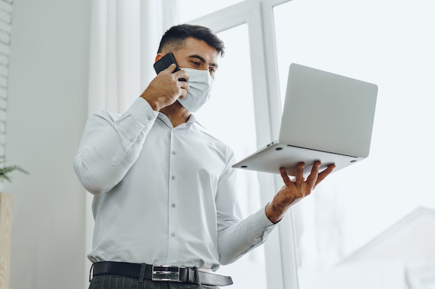 Bel homme homme d'affaires en masque médical à l'aide de son ordinateur portable tout en parlant sur son téléphone portable