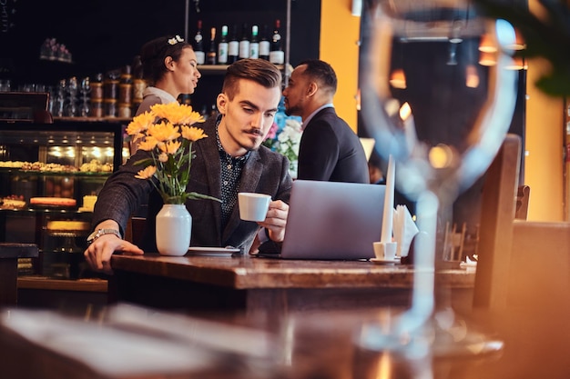 Bel homme indépendant avec une barbe et des cheveux élégants vêtus d'un costume noir travaillant sur un ordinateur portable assis dans un café.