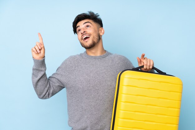 bel homme isolé sur bleu en vacances avec valise de voyage et pointant vers le haut