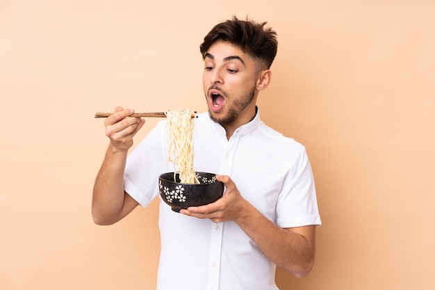 Bel homme isolé sur un mur beige tenant un bol de nouilles avec des baguettes et le manger