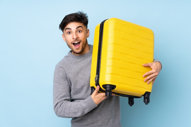 Bel homme isolé sur mur bleu en vacances avec valise de voyage et surpris