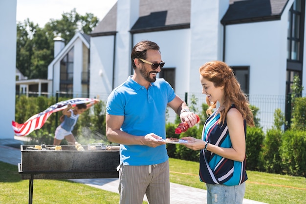 Bel homme joyeux souriant à sa femme tout en mettant du ketchup dans son assiette