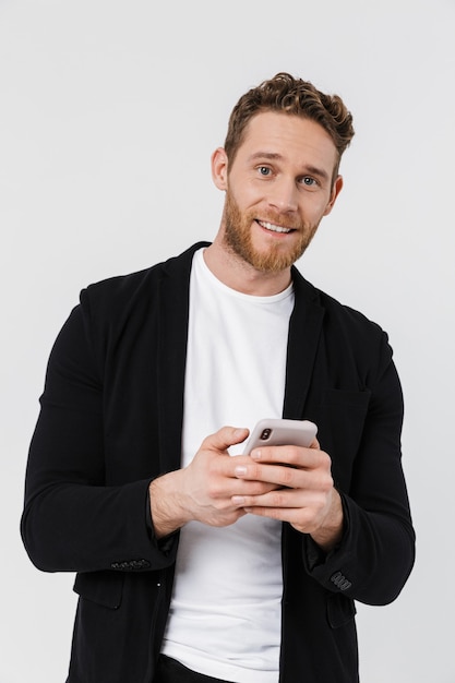 bel homme joyeux en veste souriant et utilisant un smartphone isolé sur un mur blanc