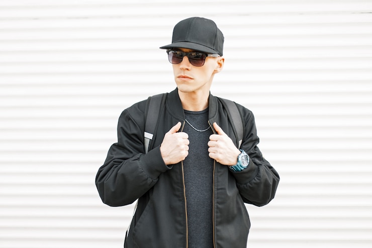 Photo bel homme avec des lunettes noires, une casquette, une veste avec un sac à dos près de la paroi métallique