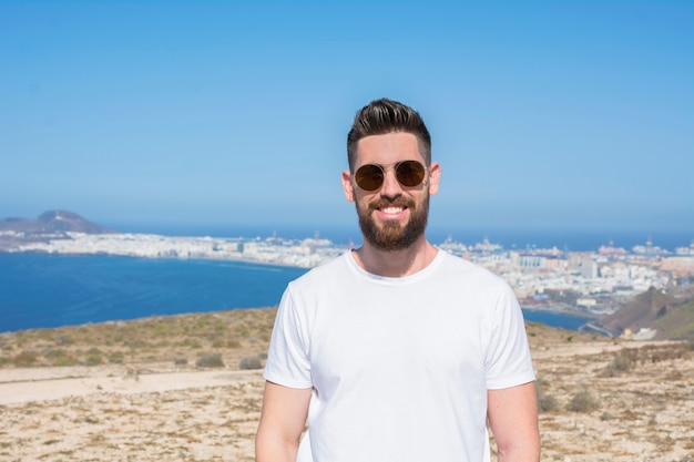 Bel homme avec des lunettes de soleil, une barbe et une chemise blanche au sommet de la montagne