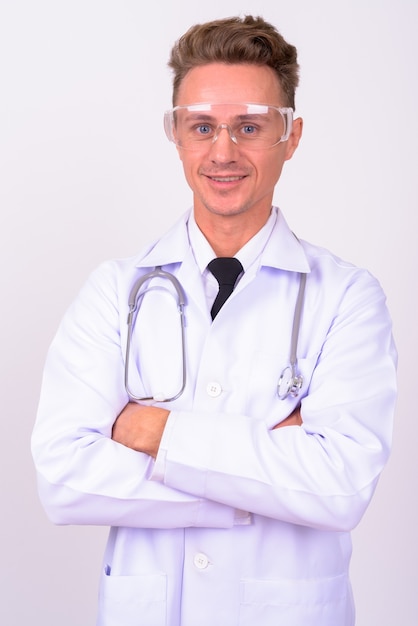 Bel homme médecin aux cheveux blonds bouclés portant des lunettes de protection contre le mur blanc