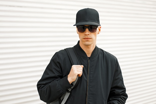 Bel homme à la mode avec des lunettes noires avec des vêtements modernes noirs près du mur blanc