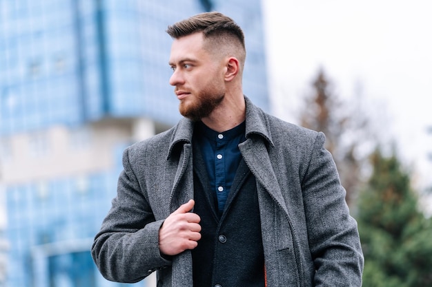 Bel homme à la mode en manteau gris et costume sombre. Arrière-plan flou de la ville d'automne.