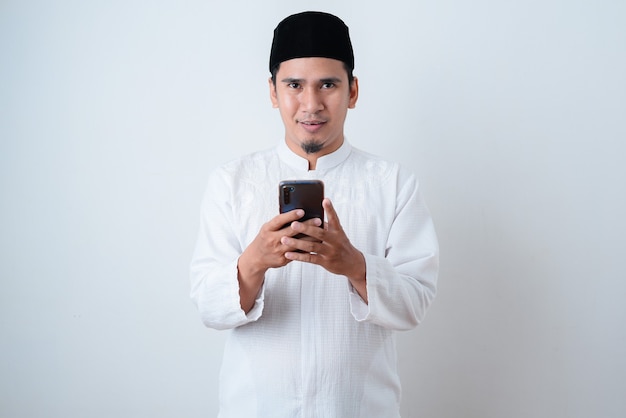 Bel homme musulman portant des vêtements musulmans et tenant son téléphone en regardant la caméra contre le mur blanc