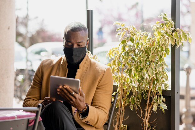 Bel homme noir en costume élégant et élégant assis dans une table basse à l'aide d'une tablette. Guy portant un masque facial.