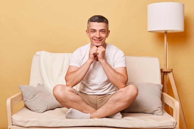 Bel homme optimiste positif portant un t-shirt blanc et un short assis sur un canapé sur fond beige regardant cmaera avec une expression ravissante visage heureux