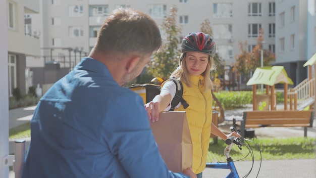 Bel homme ouvre la porte à une femme de livraison de nourriture portant un sac à dos thermique jaune sur un vélo et reçoit une commande ou un colis avec de la nourriture Concept de livraison de nourriture