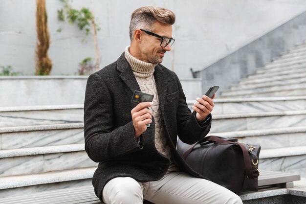 Bel homme portant une veste shopping en ligne avec téléphone mobile et carte de crédit alors qu'il était assis à l'extérieur
