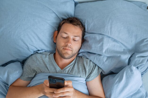 Un bel homme qui utilise son téléphone portable au lit le matin, surfe sur Internet, vérifie les réseaux sociaux et