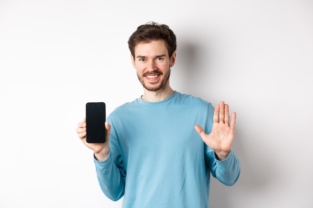 Bel homme de race blanche avec barbe, montrant un écran de smartphone vide et le numéro cinq, levant la main pour saluer et dire bonjour, debout sur fond blanc.