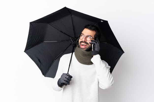 Bel homme de race blanche avec barbe tenant un parapluie sur un mur blanc isolé, gardant une conversation avec le téléphone mobile