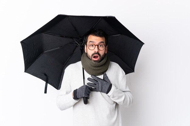 Bel homme de race blanche avec barbe tenant un parapluie sur un mur blanc isolé surpris et choqué tout en regardant à droite