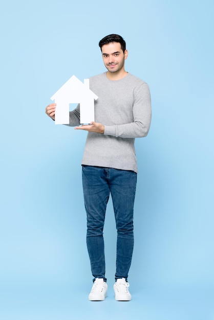 Bel homme de race blanche souriant tenant la découpe du modèle de maison debout dans un fond de studio isolé bleu clair