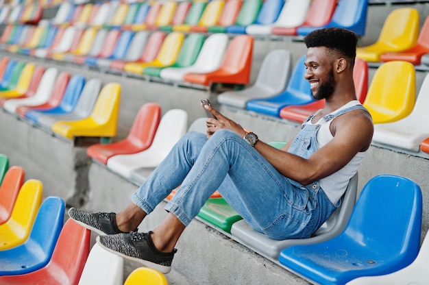 Bel homme à une salopette en jean avec téléphone portable aux mains posé sur des chaises colorées au stade Portrait d'homme noir à la mode.