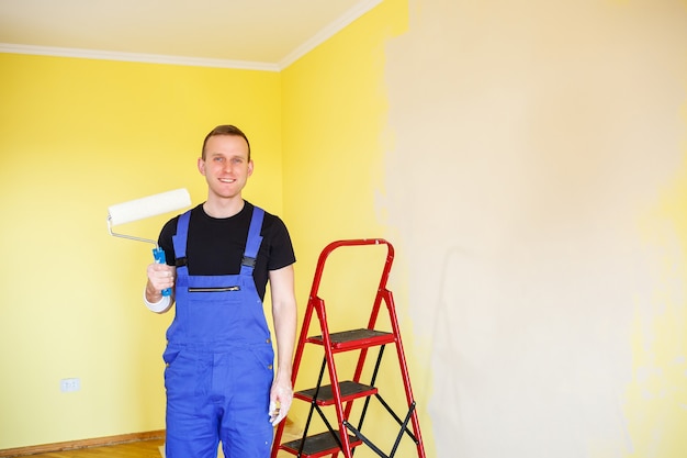 Un bel homme en salopette tient un rouleau à peinture. Accessoires d'outils pour la rénovation d'une pièce d'appartement. Concept de rénovation domiciliaire