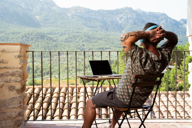 bel homme se détendre et travailler sur un ordinateur portable sur le balcon de sa maison, tout en écoutant de la musique et en profitant de la vue sur la montagne
