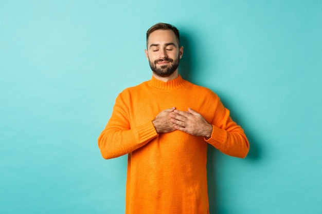 Bel homme se sentant nostalgique, se tenant la main sur le cœur et rêvassant, se souvenant ou imaginant quelque chose, debout insouciant sur fond turquoise.