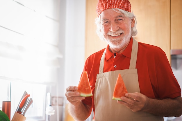 Bel homme senior caucasien dans la cuisine de la maison tout en coupant des tranches de pastèque rouge fraîche retraité barbu souriant actif vêtu de rouge avec un bandana sur la tête