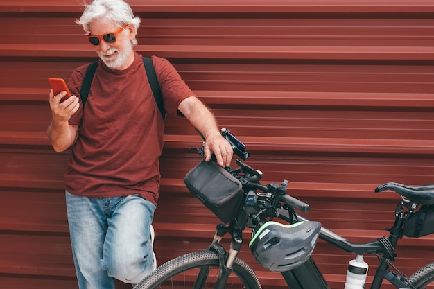 Bel homme senior debout près de son vélo électrique dans la ville portant des lunettes de soleil à l'aide de téléphone