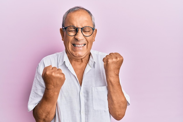 Bel homme senior mature portant une chemise décontractée et des lunettes célébrant la surprise et l'émerveillement du succès avec les bras levés et les yeux fermés