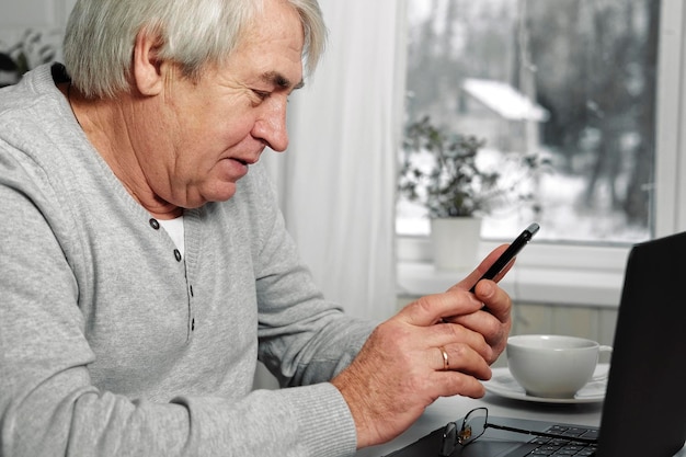 Bel homme senior sérieux portant des lunettes à l'aide d'un téléphone portable tout en étant assis à son bureau sur son lieu de travail confortable avec un ordinateur portable