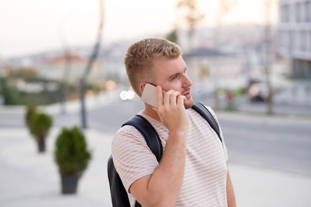 Bel homme sérieux barbu blond portant un sac à dosvêtements décontractés ayant un appel à l'aide d'un smartphone dans la ville à l'extérieurUn mec caucasien portant un sac à dos parlant par téléphone