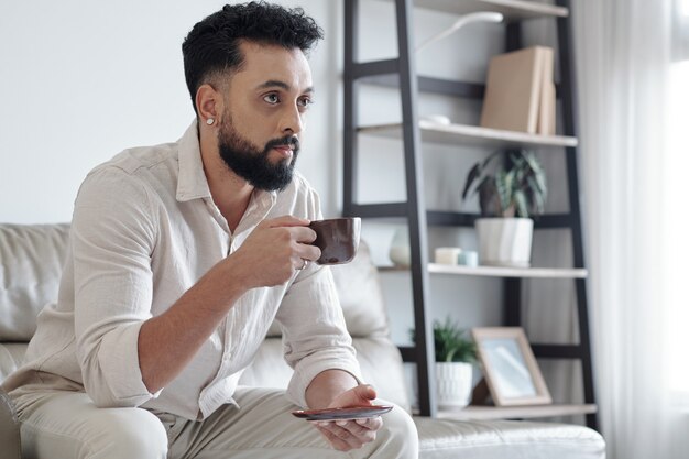 Bel homme sérieux buvant une tasse d'espresso et regardant des nouvelles ou une émission à la télévision à la maison