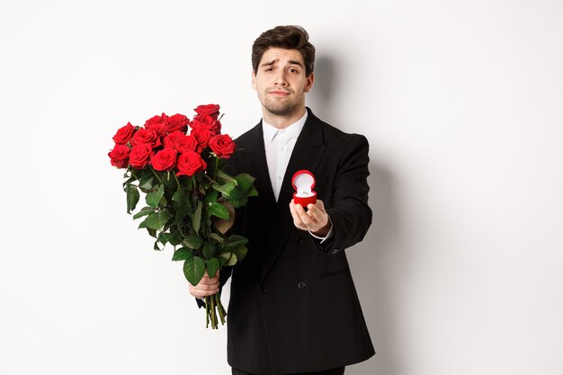 Bel homme souriant en costume noir, tenant des roses et une bague de fiançailles, faisant une proposition pour l'épouser, debout sur fond blanc