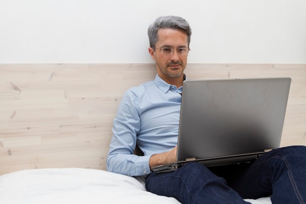 Le bel homme souriant avec des lunettes aux cheveux gris assis sur le lit et travaillant sur un ordinateur portable.