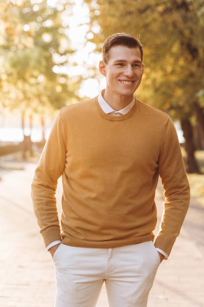 Bel homme souriant moderne en vêtements jaunes et blancs posant dans le parc au coucher du soleil