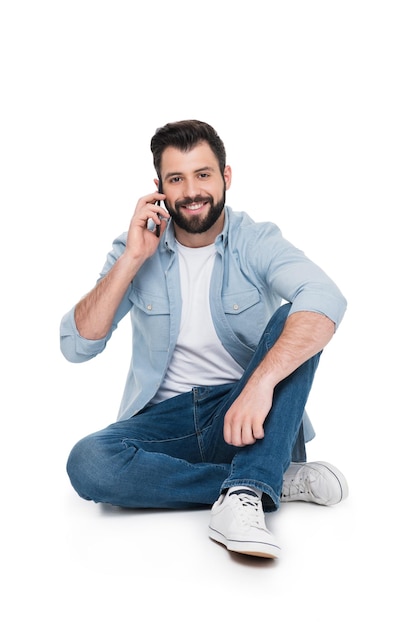 Bel homme souriant parlant sur smartphone tout en étant assis isolé sur blanc