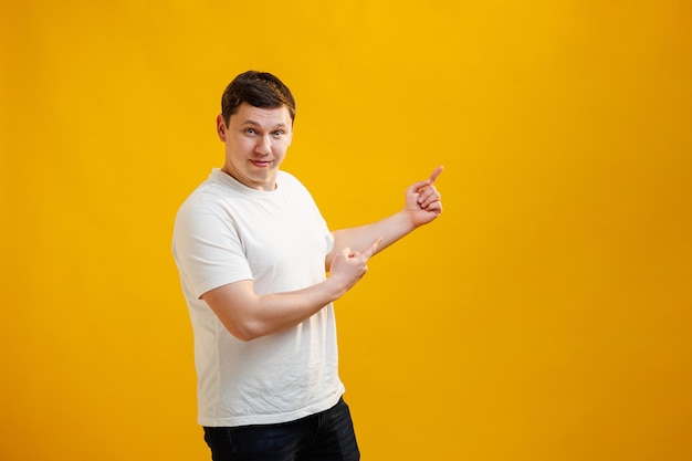 Bel homme souriant portant un t-shirt blanc debout pointant les doigts de côté sur l'espace de copie maquette sur fond de studio jaune