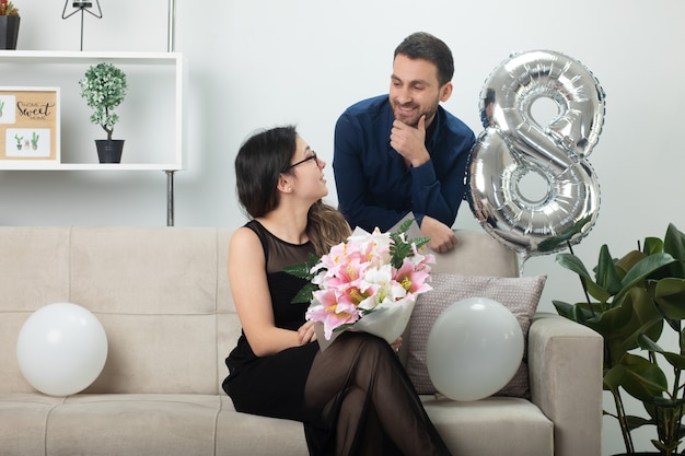 Bel homme souriant regardant une jolie jeune femme dans des lunettes optiques tenant un bouquet de fleurs assis sur un canapé dans le salon le jour de la journée internationale de la femme en mars