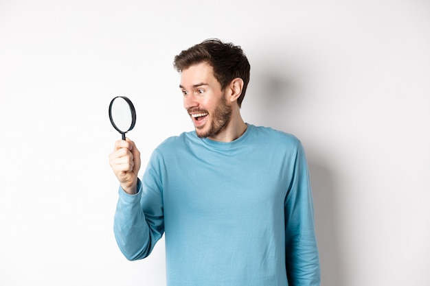 Bel homme souriant regardant à travers une loupe avec un visage étonné, a trouvé une promotion intéressante, debout sur fond blanc