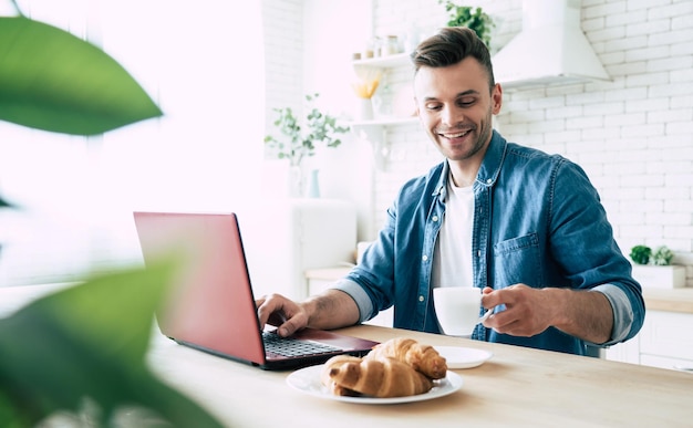 Bel homme souriant utilise un ordinateur portable et boit du café ou du thé assis dans la cuisine à la maison