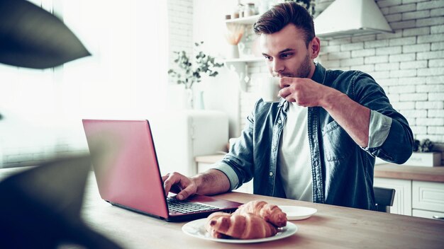 Bel homme souriant utilise un ordinateur portable et boit du café ou du thé assis dans la cuisine à la maison