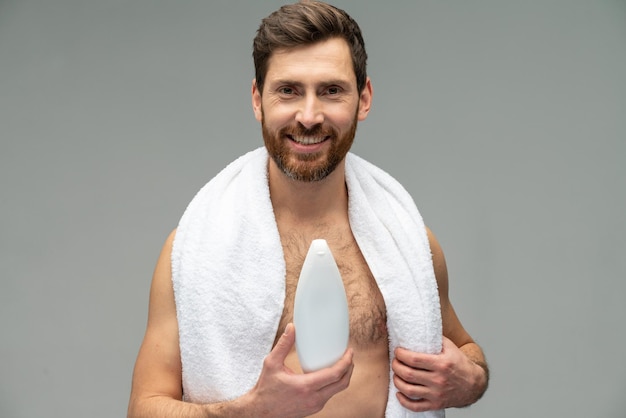 Bel homme sportif fort avec une serviette sur le cou tenant du shampoing et se préparer à la douche isolé sur fond gris studio Concept de douche