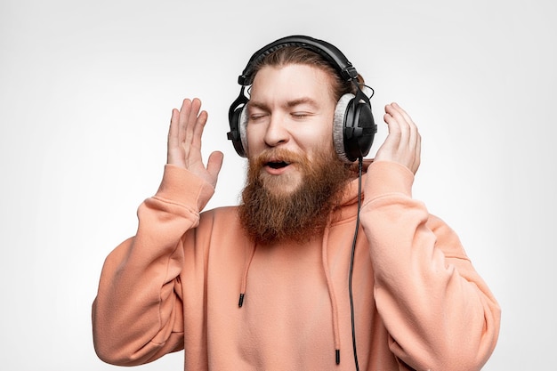 Un bel homme surpris scandinave rit et écoute de la musique dans des écouteurs professionnels isolés sur fond gris. Un mec heureux avec une coiffure au gingembre, une barbe. Technologies numériques modernes