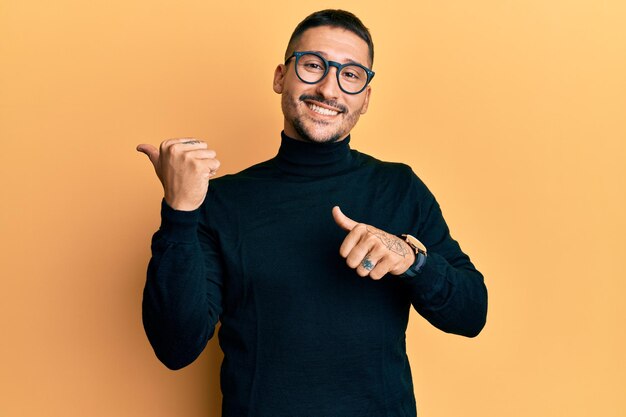 Bel homme avec des tatouages portant un pull à col roulé et des lunettes pointant vers l'arrière derrière avec la main et les pouces vers le haut souriant confiant