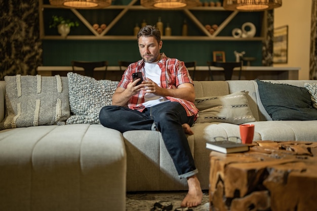 Bel homme avec téléphone portable assis à la maison homme américain à l'aide de téléphone tout en étant assis sur un canapé à liv