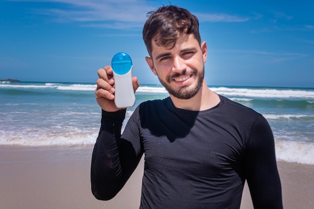 Bel homme tenant un écran solaire sur la plage souriant en regardant la caméra