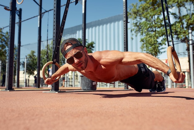 Bel homme sur un terrain de sport Le sportif le gars effectue des exercices difficiles de gymnastique sportive Homme musclé faisant de l'exercice avec des anneaux Le concept d'un jeune homme moderne